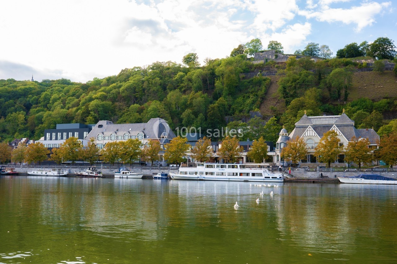 Vue sur une rivière bordée de bateaux, avec des bâtiments historiques et une colline boisée en arrière-plan.