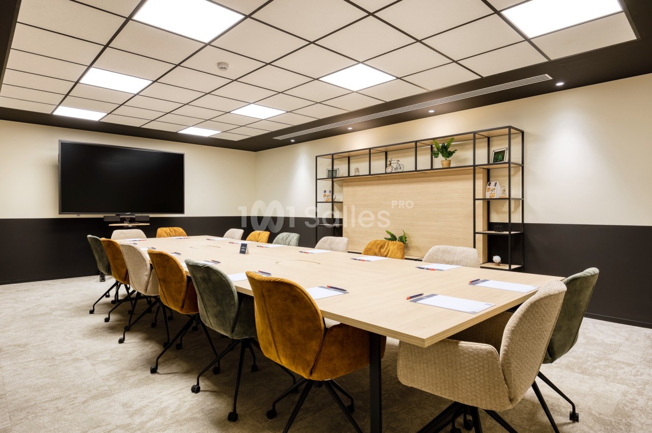 Salle de réunion moderne avec une grande table en bois, des chaises colorées et un écran mural.