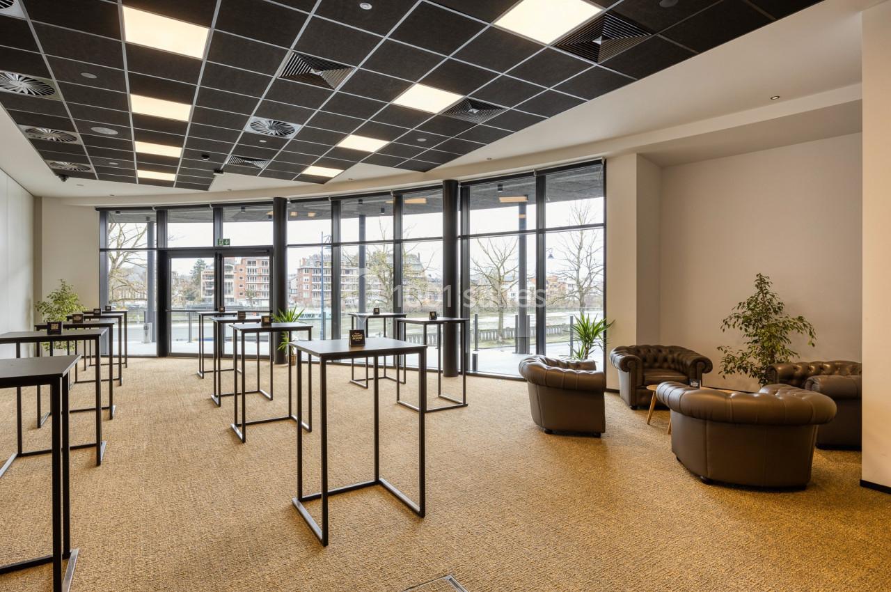 Location salle Namur (Namur) - Casino Resort Namur #15 Salle lumineuse avec grandes baies vitrées, tables hautes noires, fauteuils en cuir et plantes décoratives.
