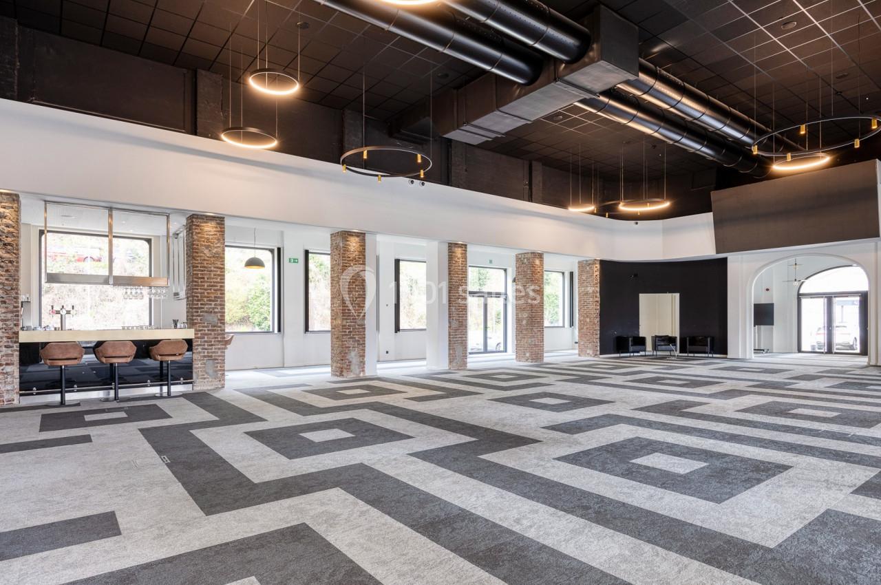 Location salle Namur (Namur) - Casino Resort Namur #20 Intérieur d'une grande salle moderne avec sol géométrique, murs en briques, bar et luminaires suspendus.