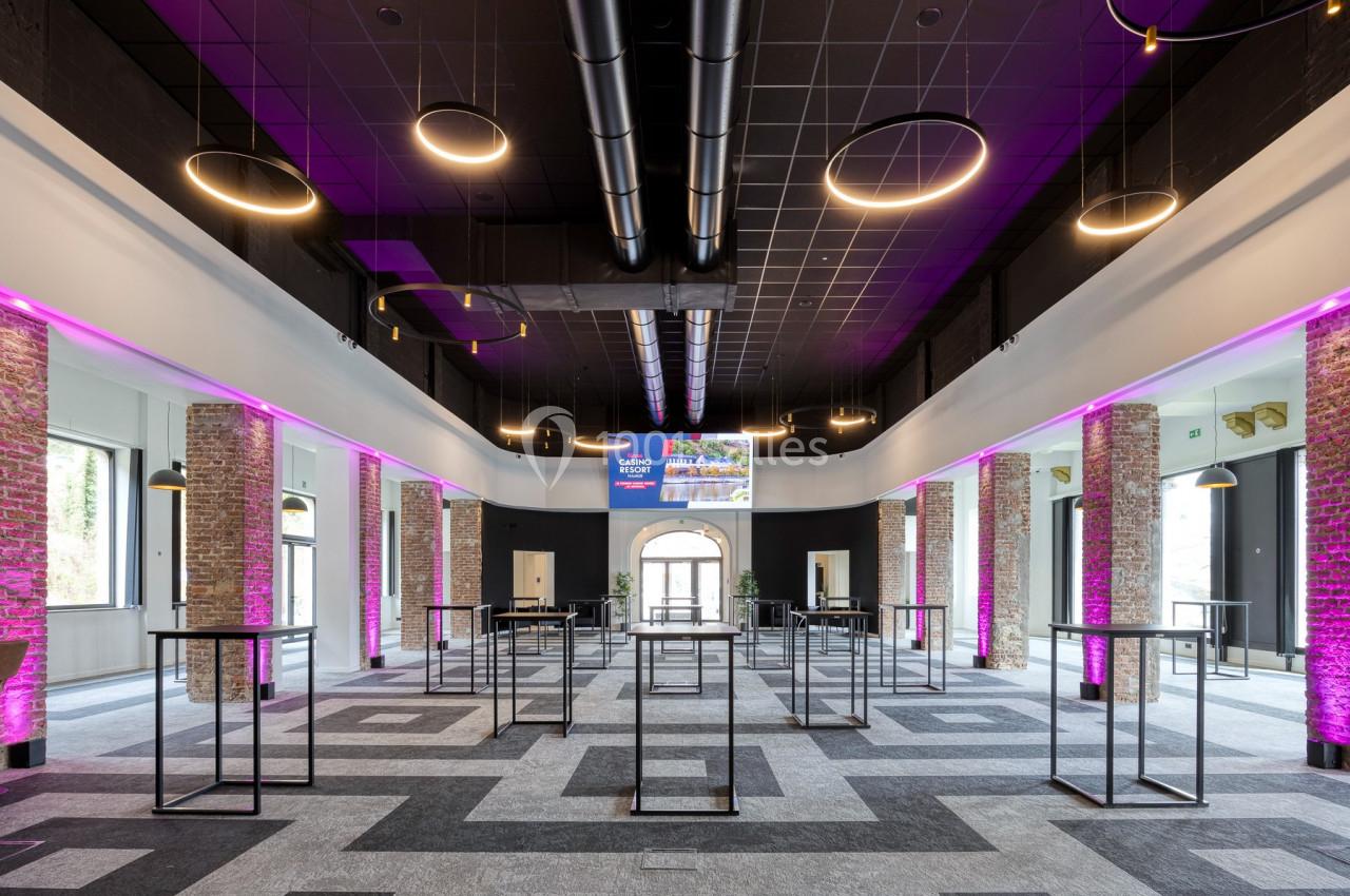 Location salle Namur (Namur) - Casino Resort Namur #25 Salle spacieuse avec murs en briques, éclairage moderne, tables hautes et lumière rose décorative.