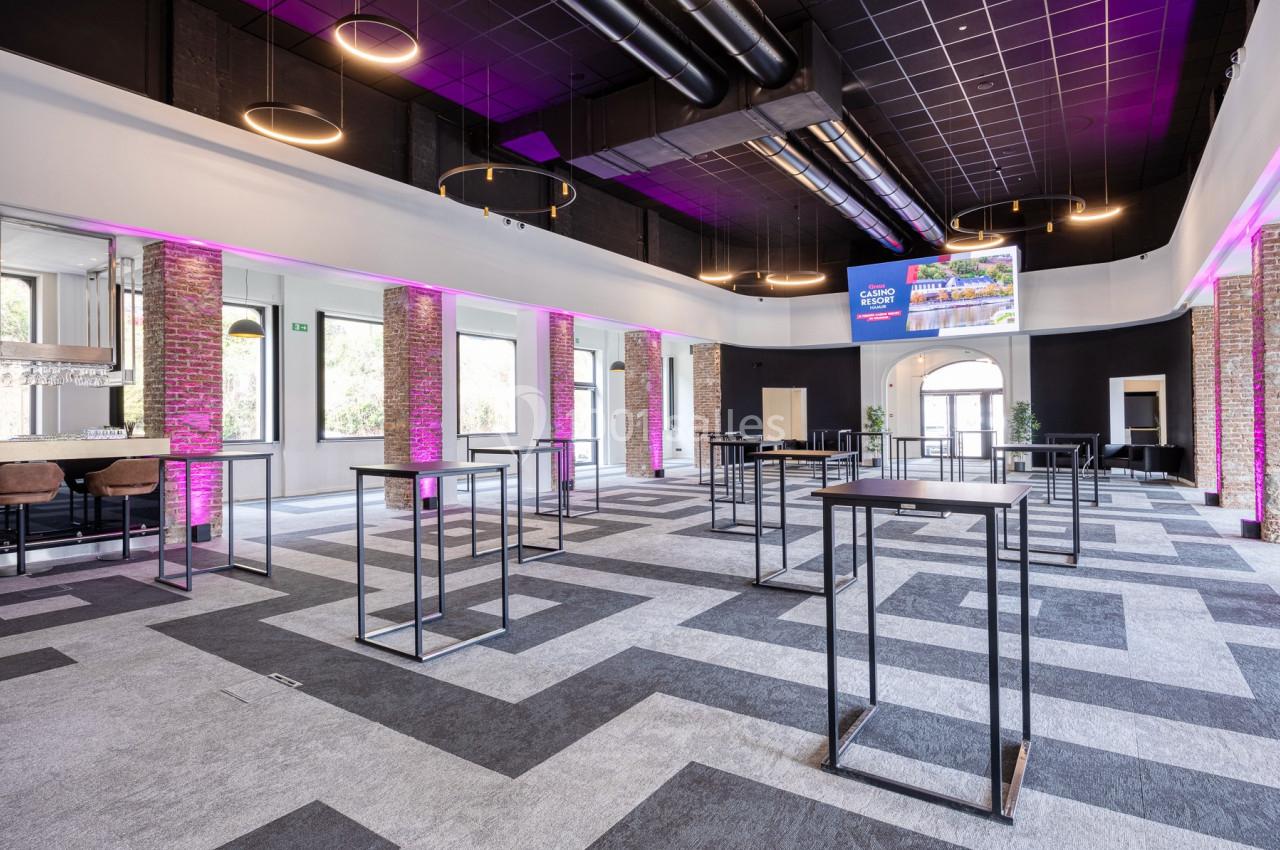 Location salle Namur (Namur) - Casino Resort Namur #28 Salle moderne avec tables hautes, éclairage suspendu, murs en briques et néons roses, sol à motifs géométriques.