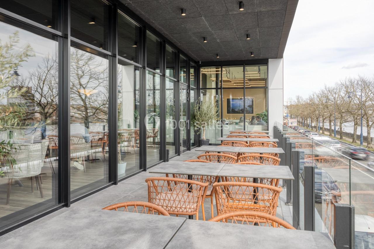 Location salle Namur (Namur) - Casino Resort Namur #34 Terrasse moderne avec tables et chaises en rotin, bordée de baies vitrées et vue sur une rue arborée.