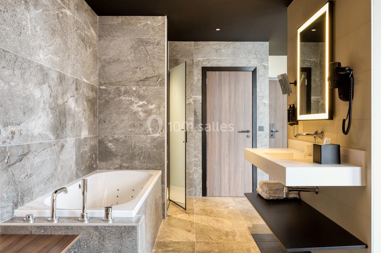 Location salle Namur (Namur) - Casino Resort Namur #47 Salle de bain moderne avec baignoire, lavabo suspendu, miroir éclairé et murs en pierre grise.