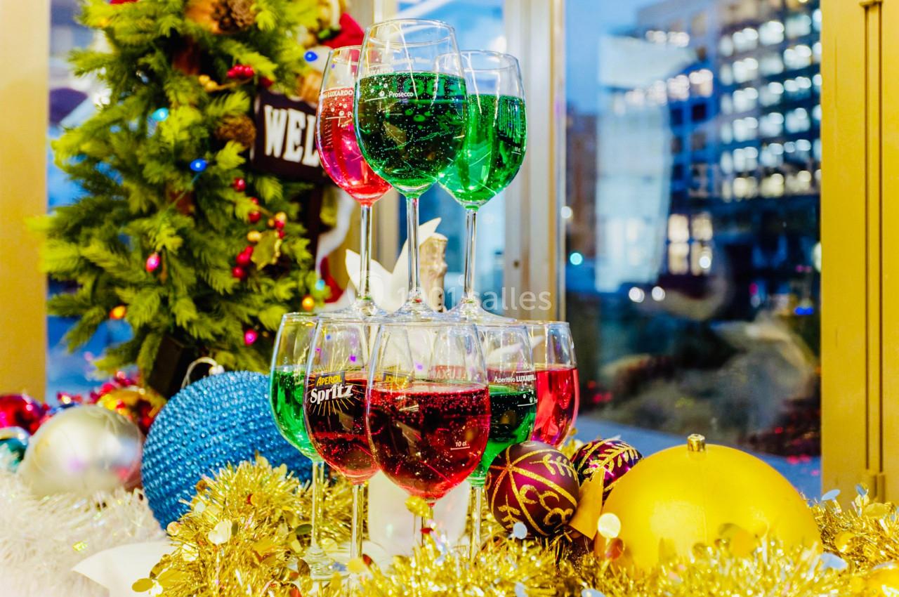 Tour de verres colorés remplis de boissons pétillantes, décorée de guirlandes et boules de Noël, près d'une fenêtre.