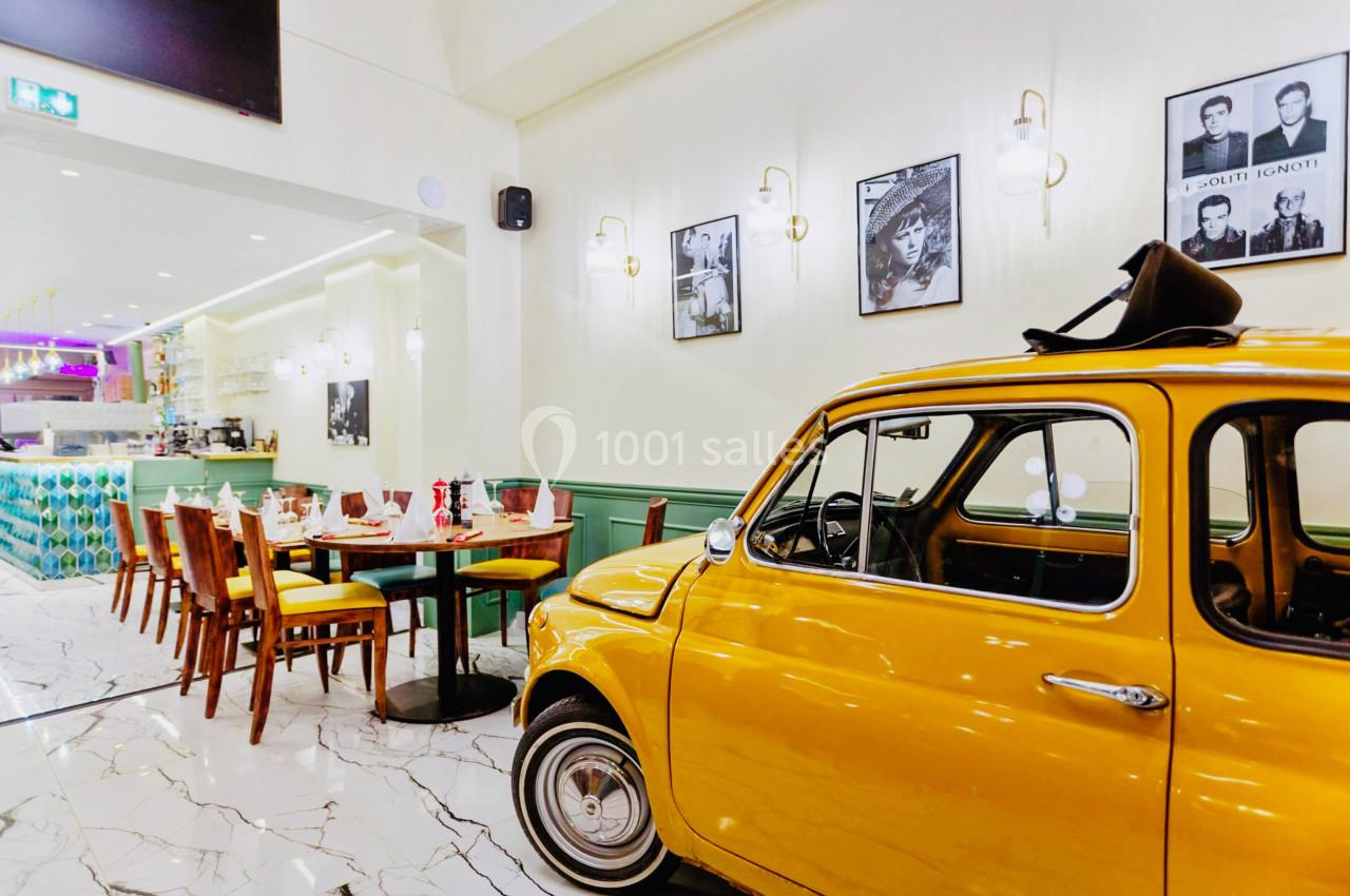 Voiture jaune vintage exposée dans un restaurant lumineux avec des tables dressées et des affiches au mur.