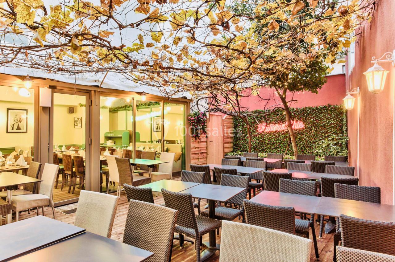 Terrasse extérieure avec tables et chaises en rotin sous une pergola végétalisée, vue sur une salle intérieure vitrée.