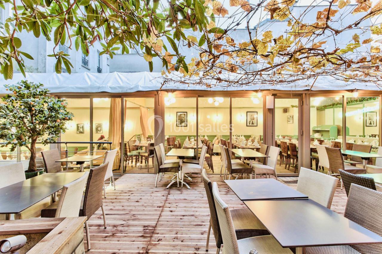 Terrasse en bois avec tables et chaises, entourée de végétation et donnant sur une salle intérieure lumineuse.