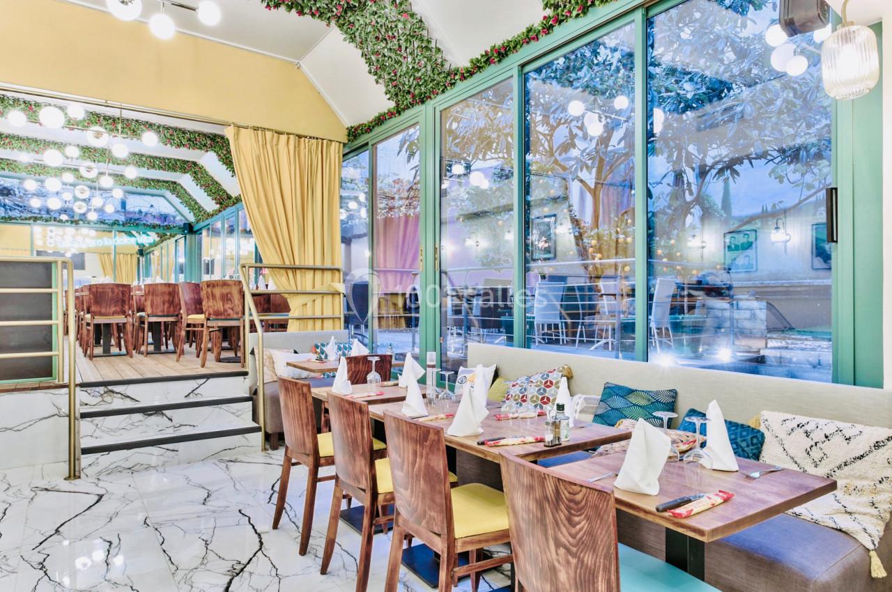 Salle de restaurant lumineuse avec tables en bois dressées, grandes baies vitrées et décoration végétale au plafond.