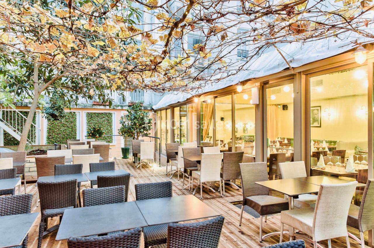 Terrasse d'un restaurant avec tables et chaises en osier, entourée de plantes et éclairée par des guirlandes lumineuses.