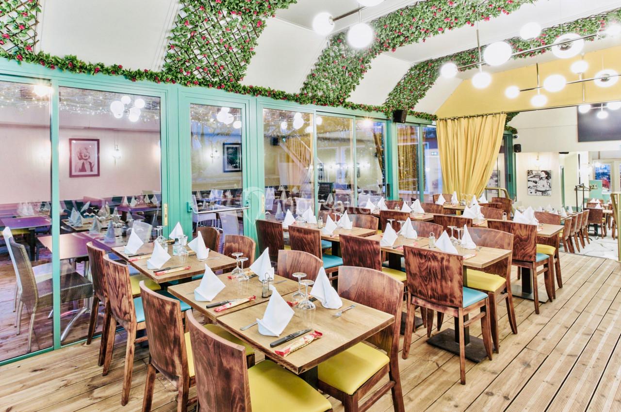 Salle de restaurant lumineuse avec tables en bois dressées, décorée de guirlandes végétales et éclairée par des suspensions…
