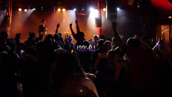 Foule dansante dans une salle sombre éclairée par des lumières colorées, avec un musicien sur scène en arrière-plan.