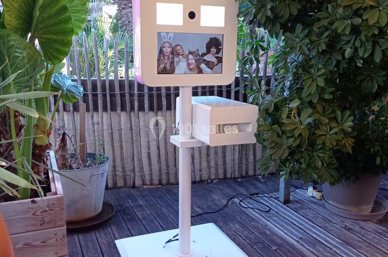 Borne photo interactive sur pied, installée en extérieur sur une terrasse en bois entourée de plantes.