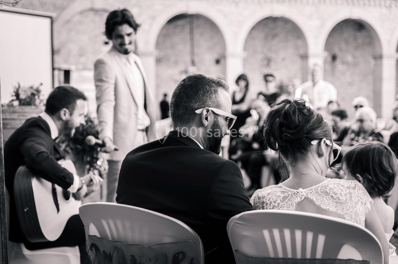 Un musicien joue de la guitare lors d'une cérémonie en plein air, avec des invités assis et un orateur debout.