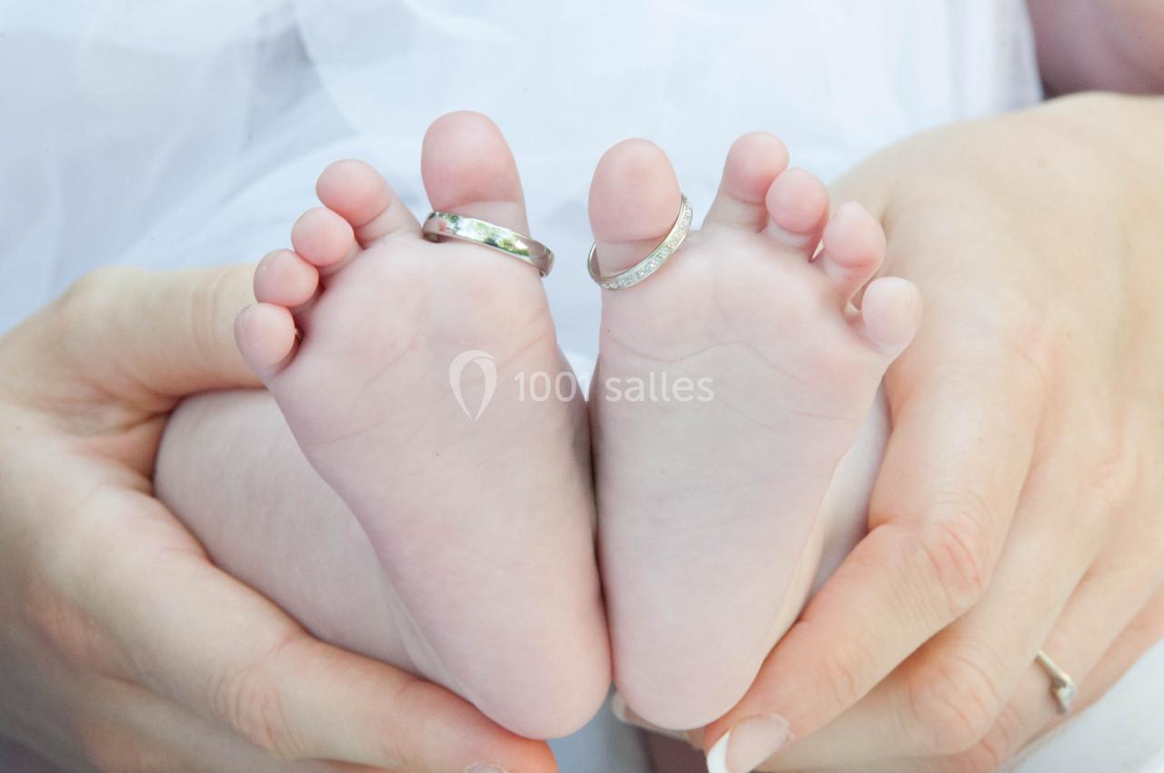 Deux pieds de bébé avec des alliances portées sur deux orteils, tenus délicatement par des mains d'adulte.