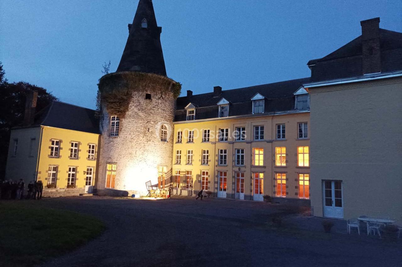 Façade éclairée d'un château avec une tour en pierre et des fenêtres illuminées à la tombée de la nuit.