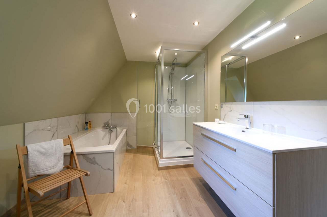 Salle de bain moderne avec baignoire, douche vitrée, meuble vasque en bois clair et chaise avec serviette.