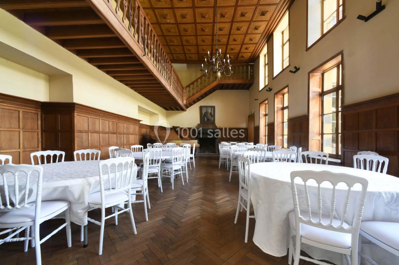 Salle lumineuse avec parquet en bois, tables rondes dressées de nappes blanches et chaises blanches, plafond à caissons.