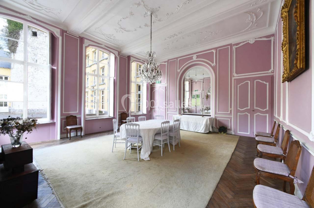 Salle à manger élégante avec murs roses, moulures blanches, lustre central, table dressée et grandes fenêtres lumineuses.