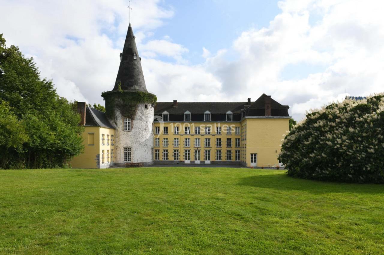 Château jaune avec une tour ronde recouverte de lierre, entouré de pelouse et d'arbres sous un ciel partiellement nuageux.