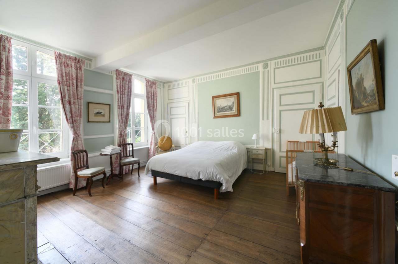 Chambre lumineuse avec lit double, parquet en bois, meubles anciens, grandes fenêtres et rideaux à motifs.