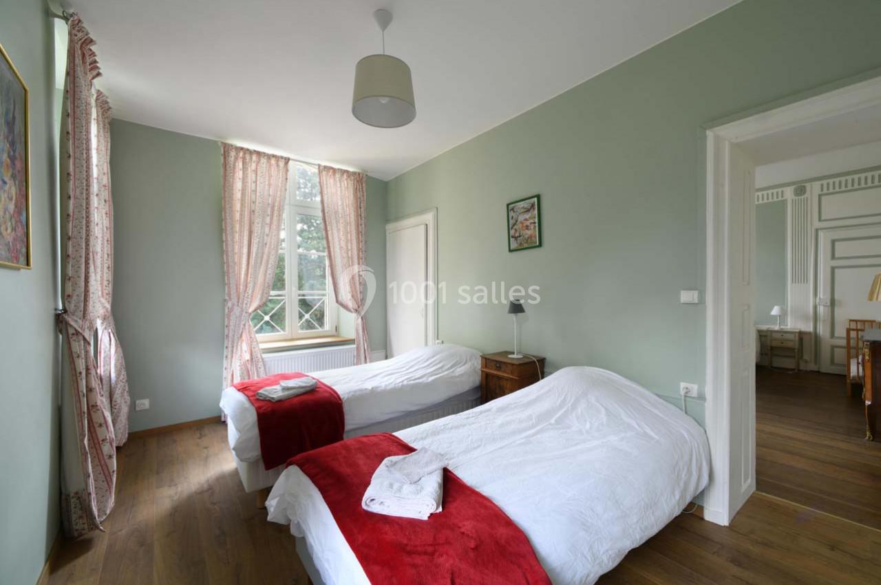 Chambre lumineuse avec deux lits simples, draps blancs, couvertures rouges, parquet et fenêtre avec rideaux roses.