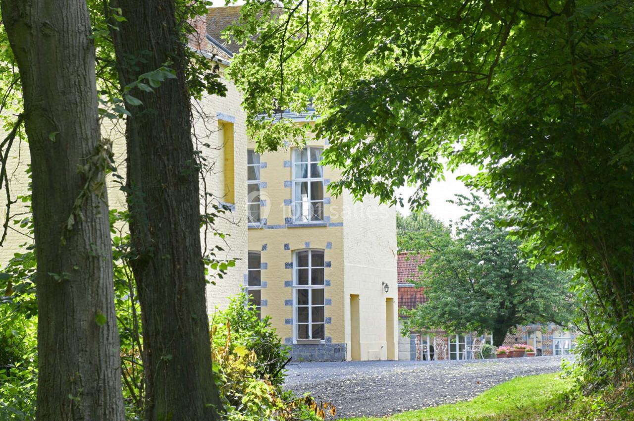 Façade d'un bâtiment en pierre claire entouré d'arbres et de végétation, avec une allée en gravier au premier plan.