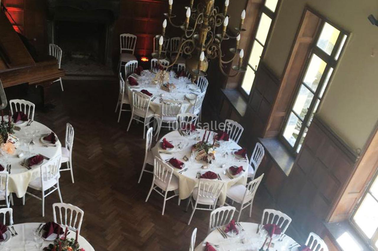 Salle de réception élégante avec tables rondes dressées, nappes blanches, serviettes rouges et chandelier central.