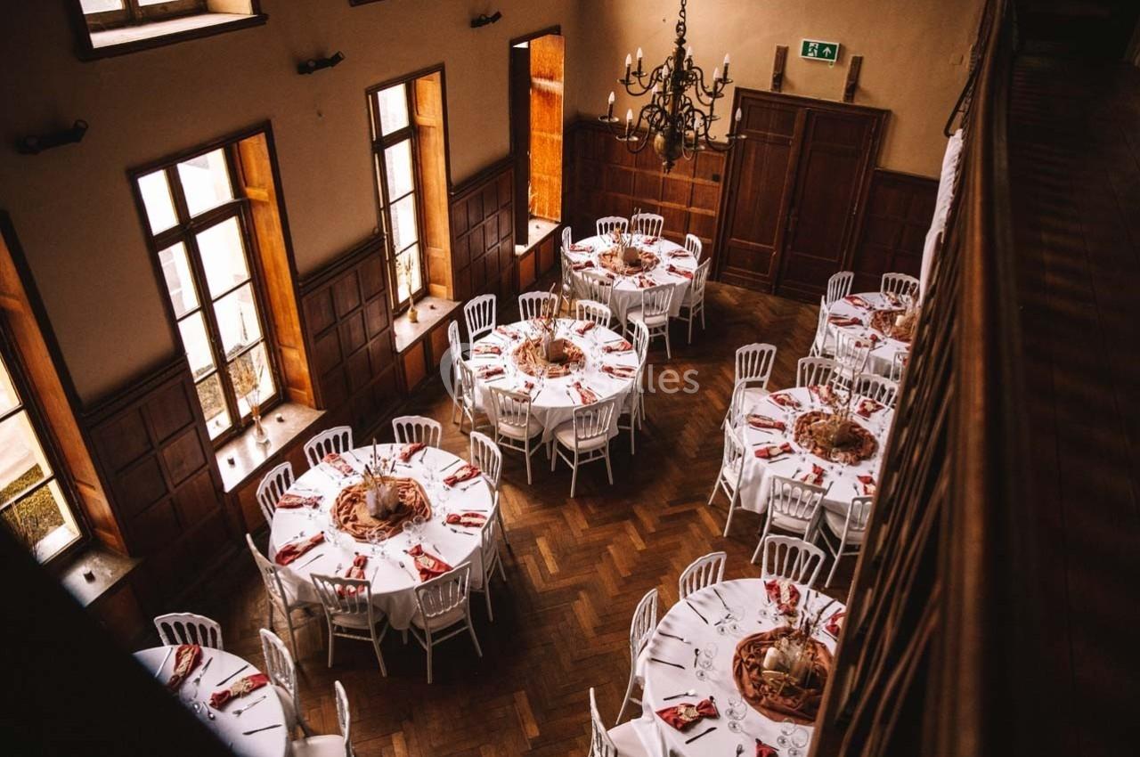 Salle de réception avec tables rondes dressées, nappes blanches, serviettes rouges et lumière naturelle entrant par les…