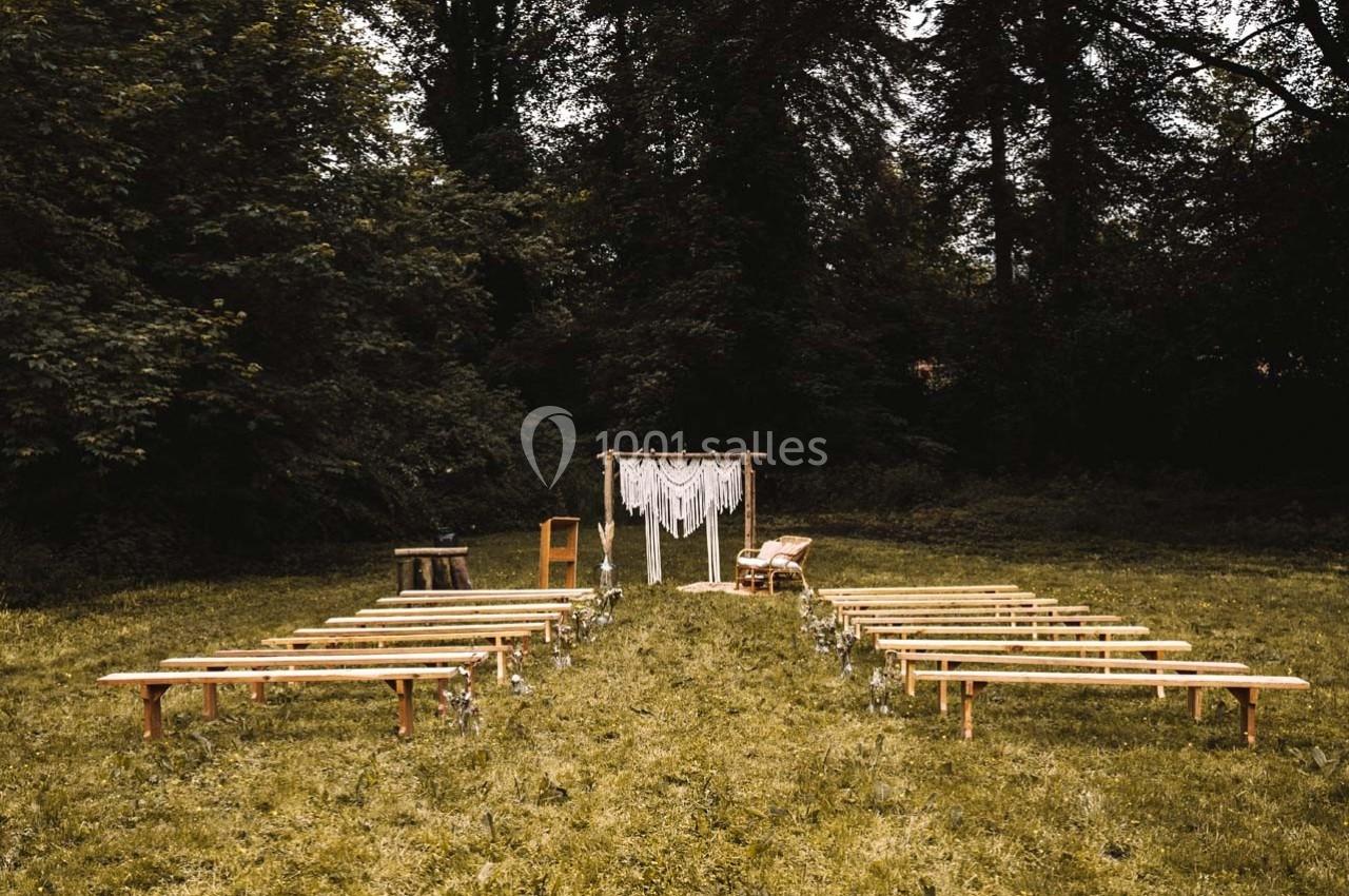 Allée centrale bordée de bancs en bois menant à une arche décorée de tissu blanc dans un cadre naturel boisé.