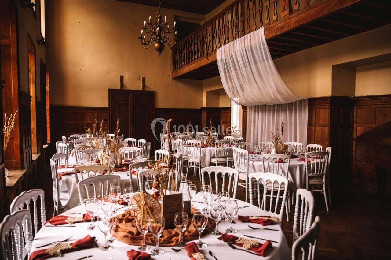 Salle de réception avec tables rondes dressées, nappes blanches, serviettes rouges et décoration champêtre.