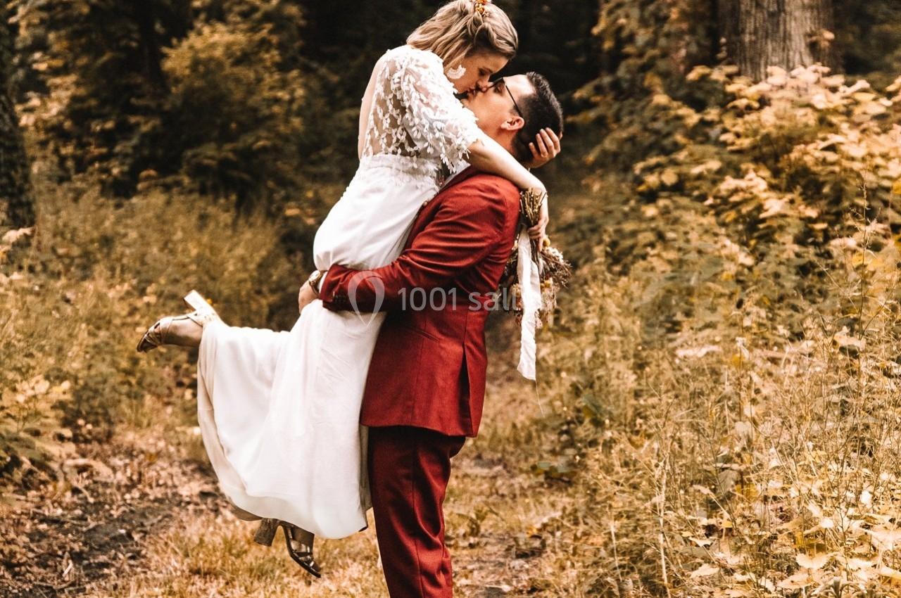 Un couple en tenue de mariage s'embrasse dans un chemin forestier, l'homme portant la femme dans ses bras.