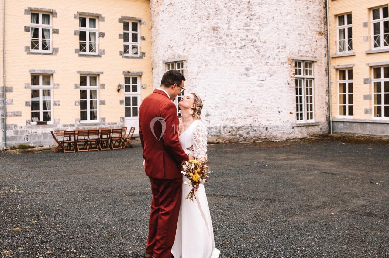 Un couple de mariés s'embrasse dans une cour pavée devant un bâtiment en pierre et en briques jaunes.