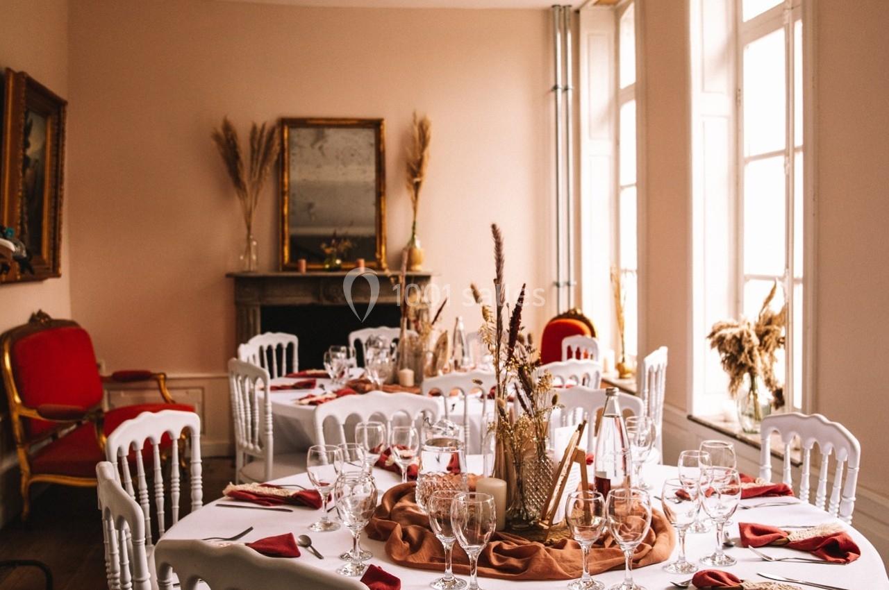 Salle de réception lumineuse avec tables dressées, décorations florales et chaises blanches autour.