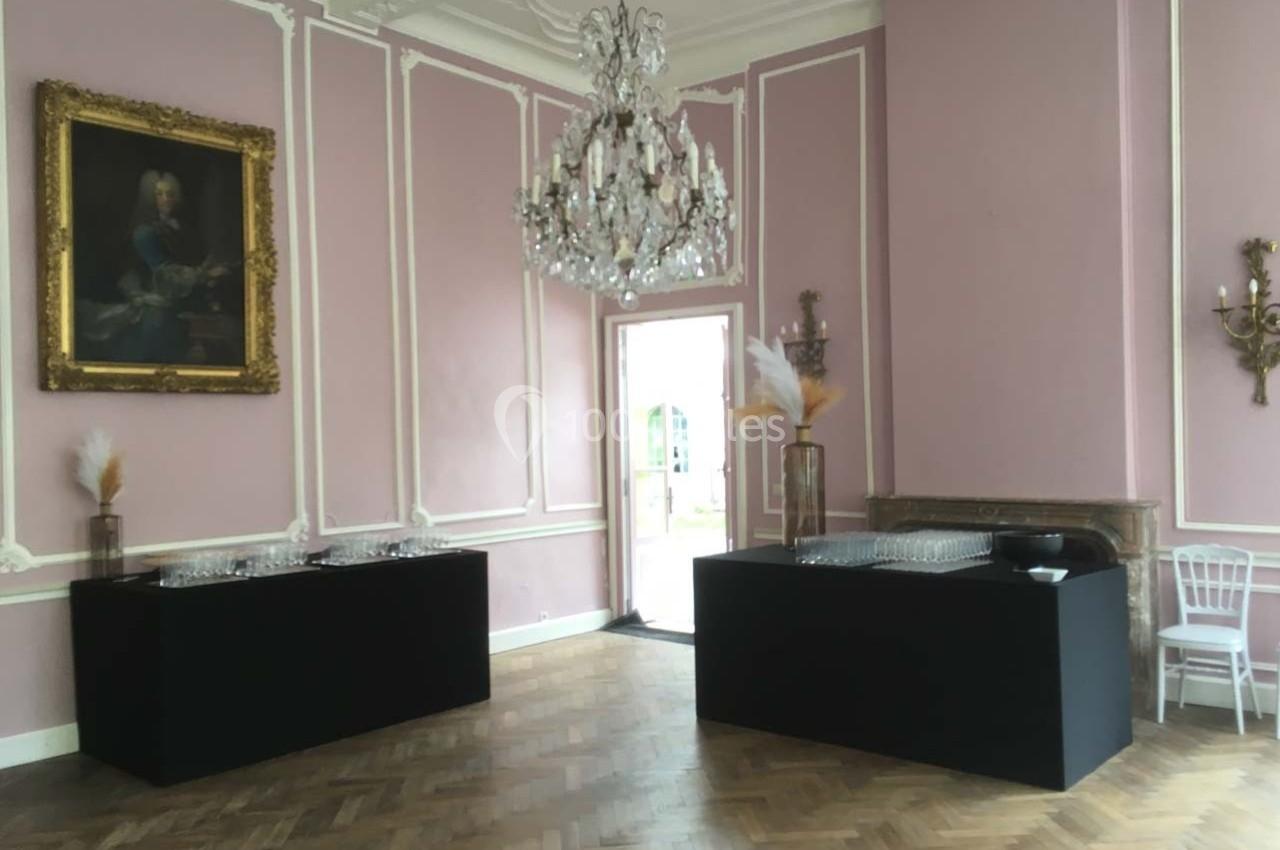 Salle élégante avec murs roses, parquet en bois, lustre en cristal, tables noires et décoration florale minimaliste.