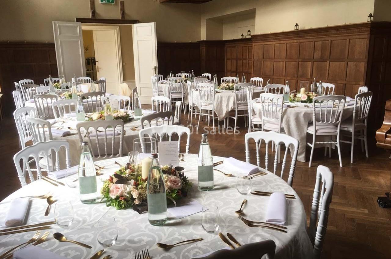 Salle de réception avec tables rondes dressées, nappes blanches, chaises blanches et décorations florales au centre.