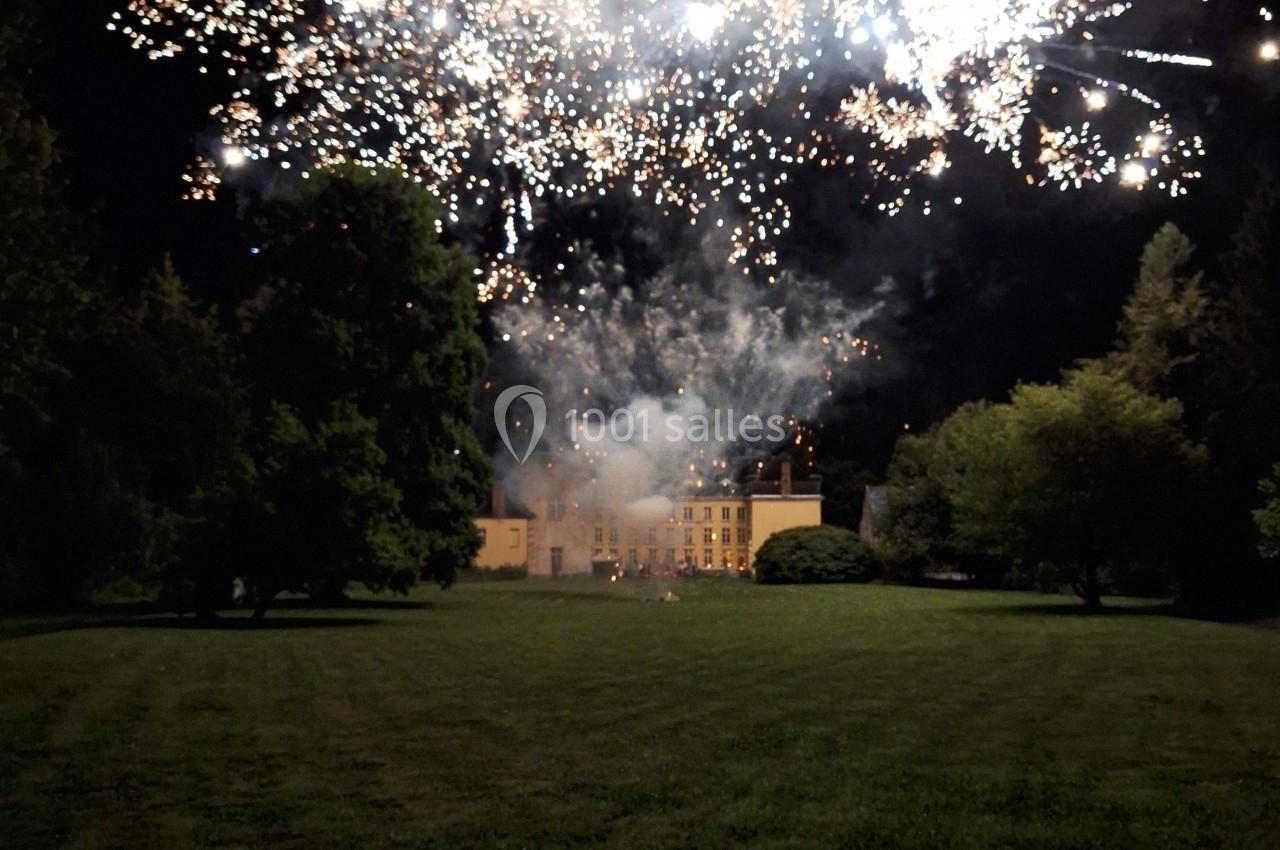 Feu d'artifice illuminant le ciel nocturne au-dessus d'un grand bâtiment entouré de pelouses et d'arbres.