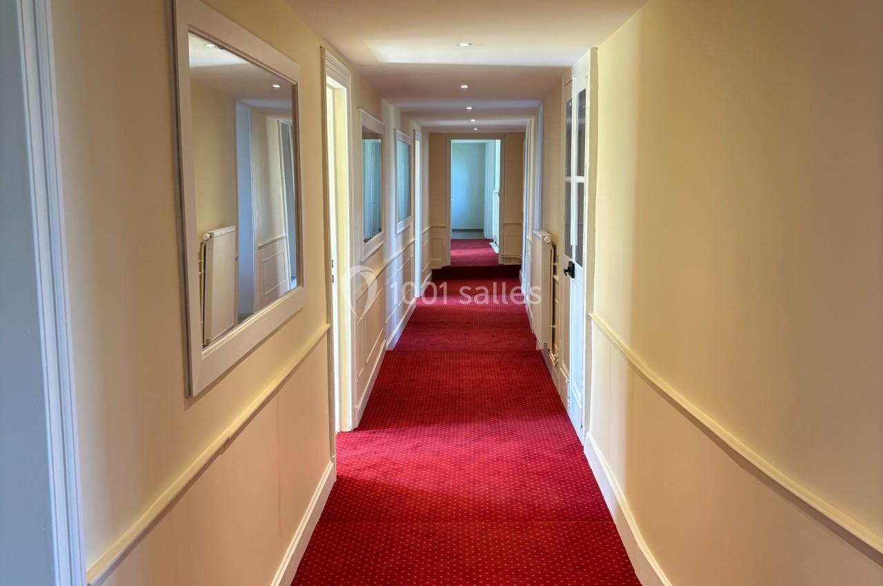 Couloir étroit avec moquette rouge, murs clairs, miroir mural et portes alignées des deux côtés.