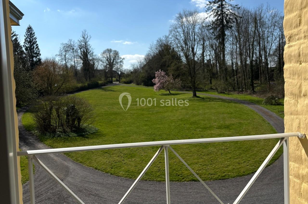 Vue depuis une fenêtre sur un jardin avec pelouse, allée sinueuse, arbres et ciel bleu en arrière-plan.