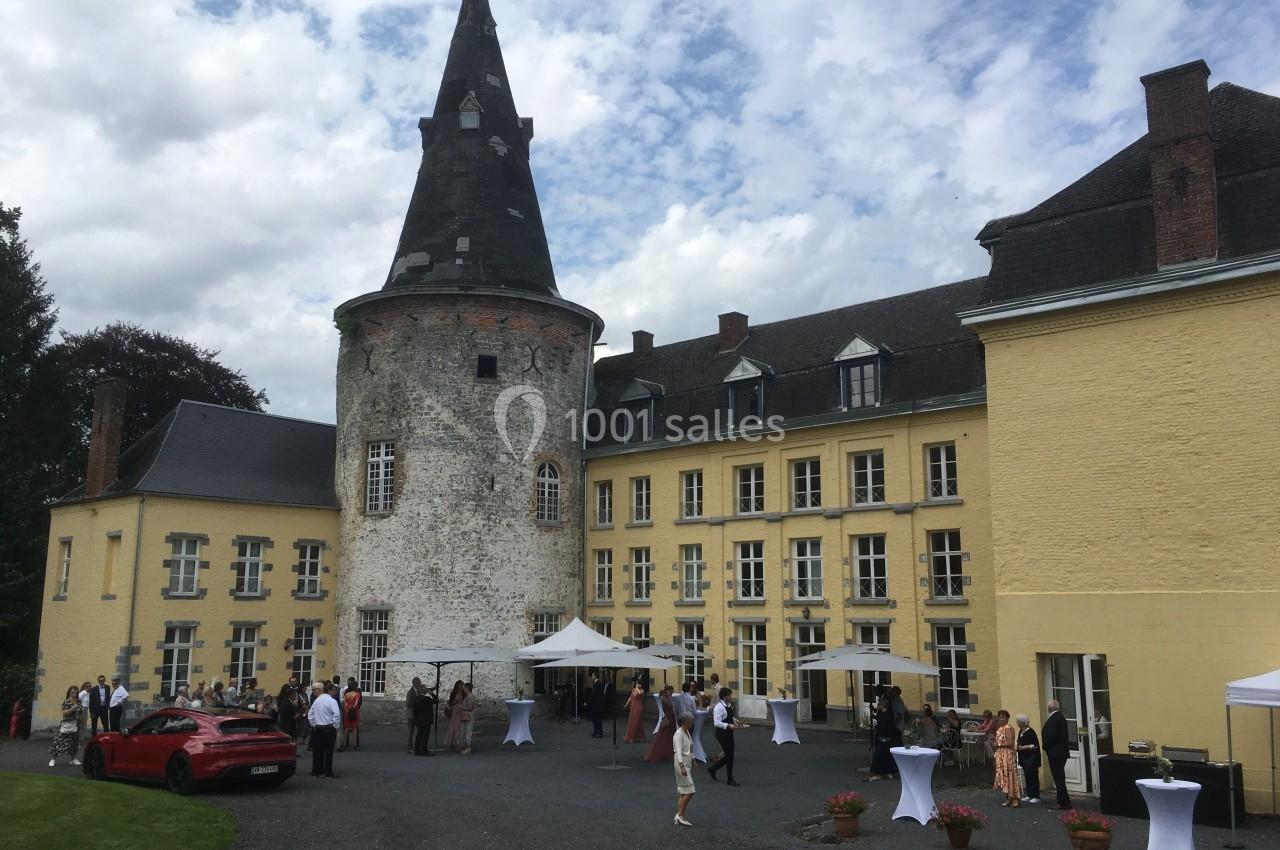 Façade d'un château avec une tour ronde, des invités rassemblés devant, tables dressées et ciel partiellement nuageux.