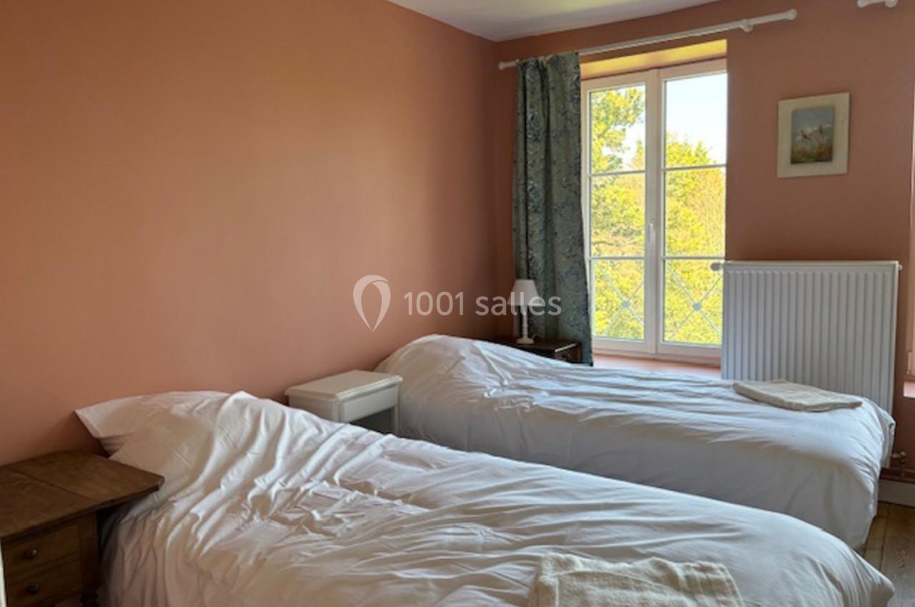 Deux lits simples avec draps blancs dans une chambre aux murs pêche, éclairée par une fenêtre donnant sur un paysage…