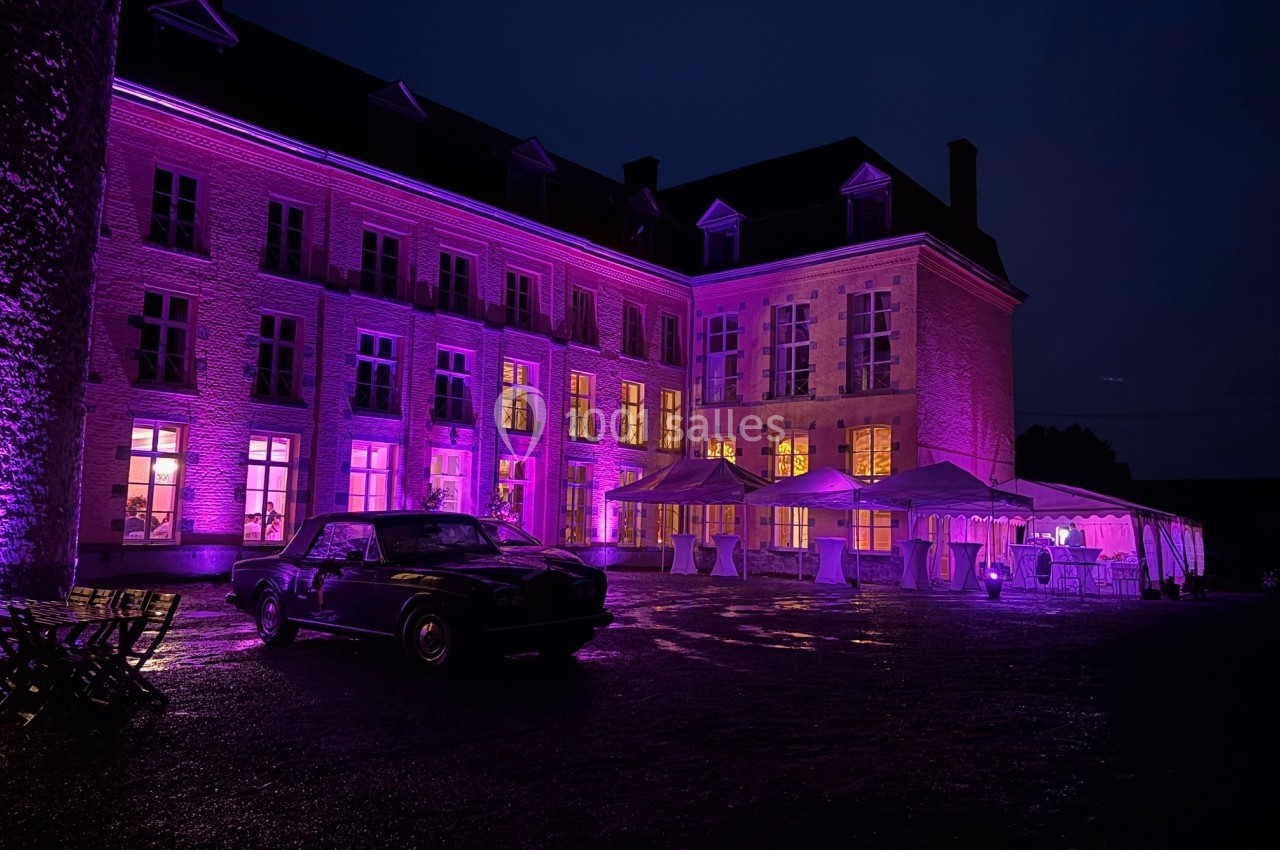 Façade d'un bâtiment éclairé en violet la nuit, avec une voiture et des tentes installées dans la cour.