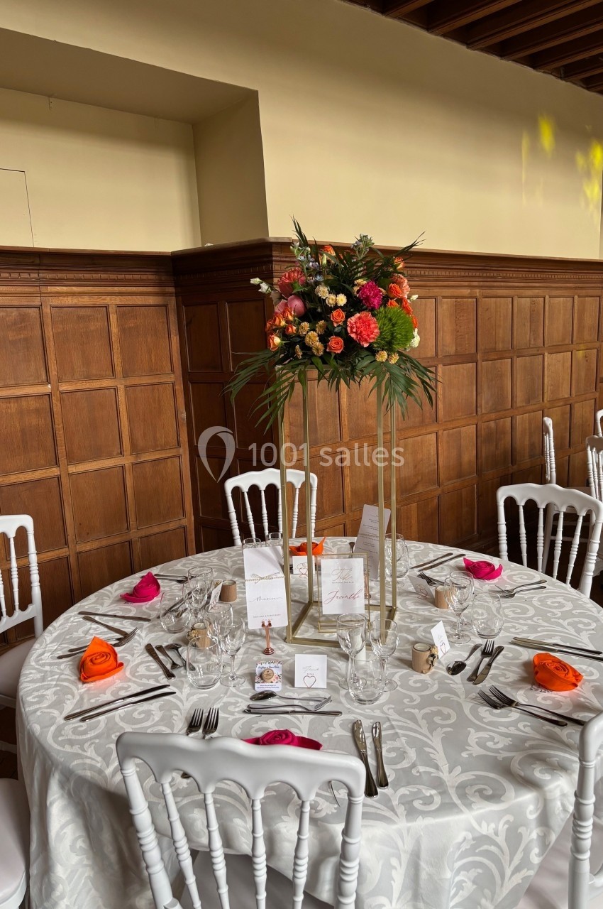 Table ronde décorée pour un événement, avec nappe blanche, chaises blanches et centre de table floral coloré.