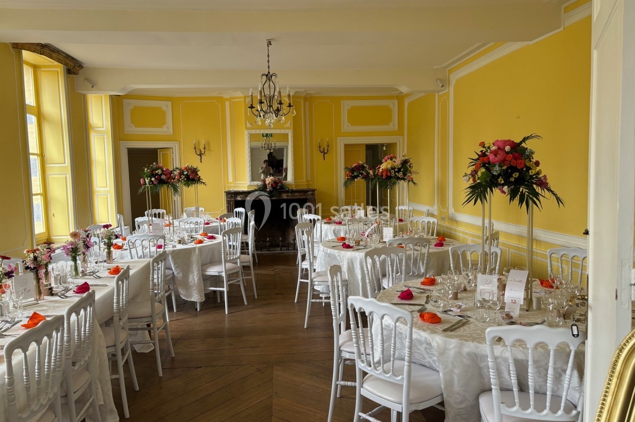 Salle de réception lumineuse avec murs jaunes, tables dressées, chaises blanches et décorations florales colorées.