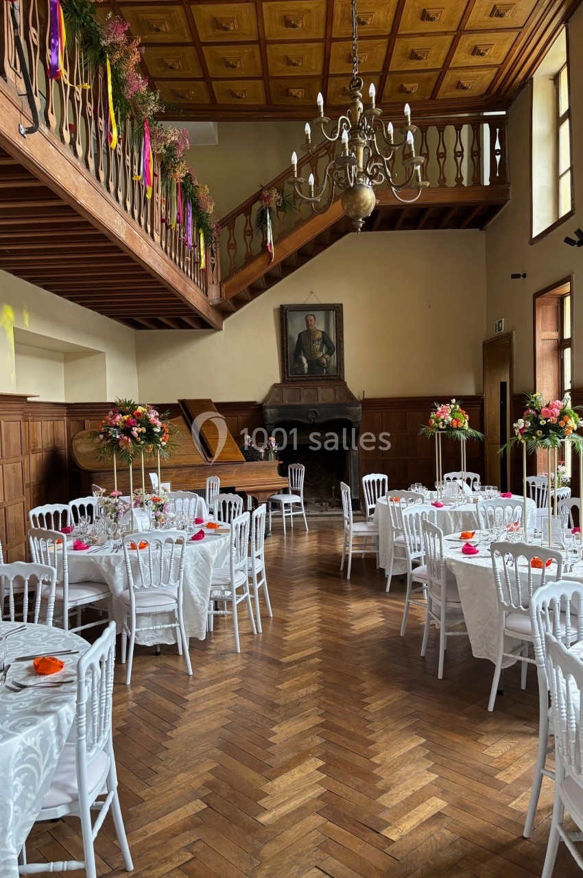 Salle de réception avec tables dressées, chaises blanches, décorations florales et un piano sous un portrait au mur.