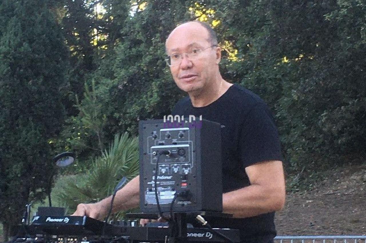 Un DJ mixe en plein air sur une table de mixage, entouré de végétation et de barrières métalliques.