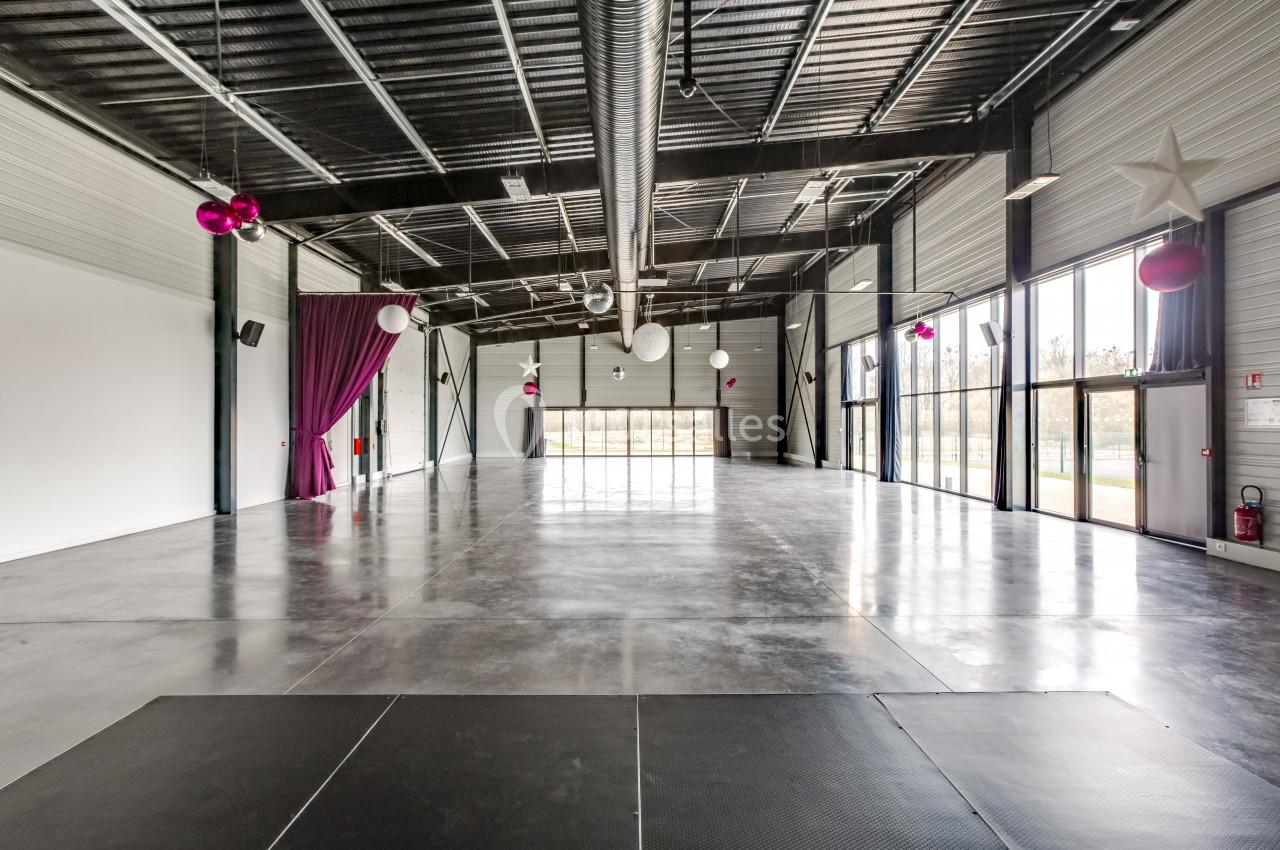 Salle spacieuse avec sol en béton, murs clairs, rideaux violets, décorations suspendues et grandes baies vitrées.