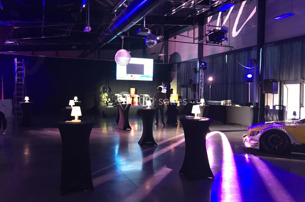 Salle événementielle avec éclairage tamisé, tables hautes éclairées et une voiture de course exposée à droite.