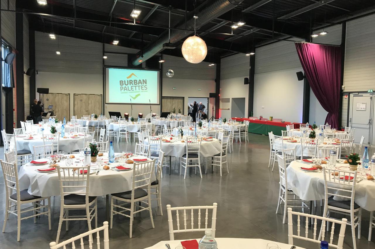 Salle de réception aménagée avec des tables rondes dressées, des chaises blanches et un écran de projection.