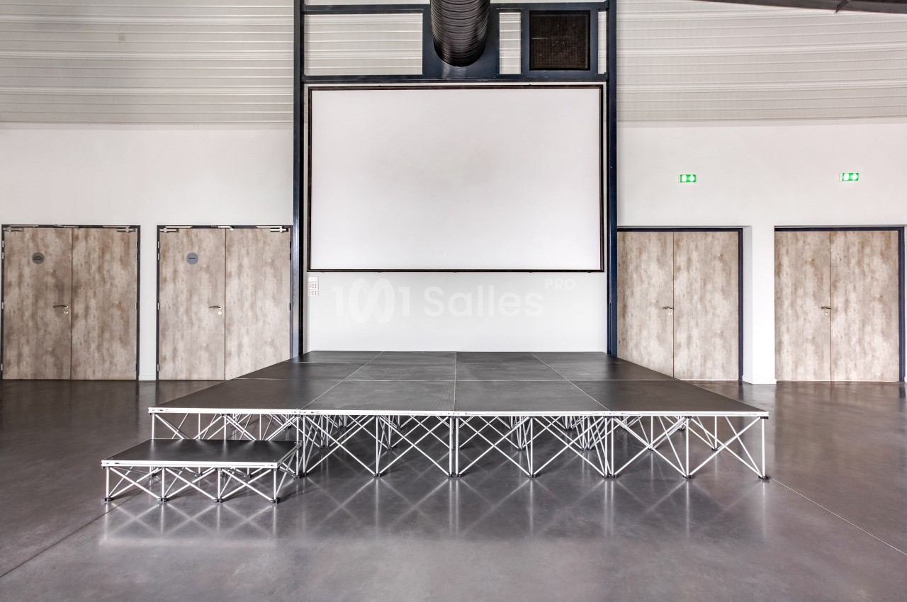 Estrade modulaire en métal avec une petite marche, installée dans une salle avec écran blanc et portes en bois.